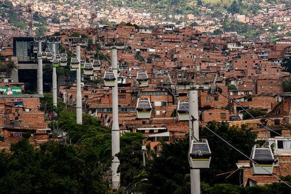 A line of cable cars floating on a zip line in the sky. Below them is an aerial view of a city with densely packed brick buildings. 