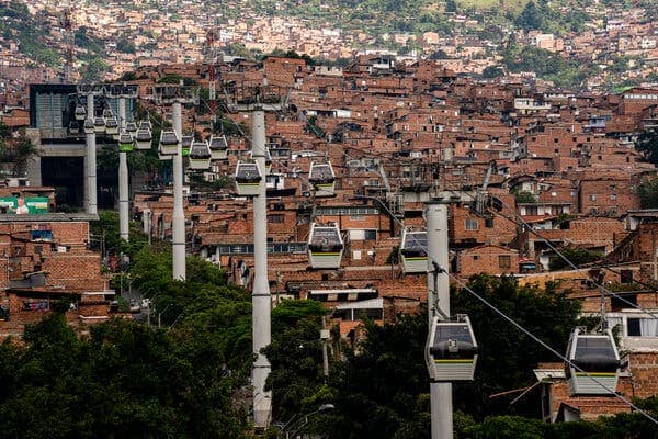 A line of cable cars floating on a zip line in the sky. Below them is an aerial view of a city with densely packed brick buildings.