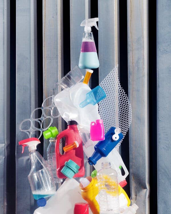 A conceptual photograph showing a spray bottle containing blue and purple liquid atop tower of laundry and cleaning containers, pill bottles, caps and other plastic material.